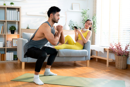 Young asian man doing squat exercise on yoga mat in living room at home while woman using phone for relax on sofa, fitness training and healthy lifestyle in a cozy, strength and strong of muscle.の写真素材