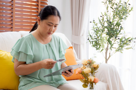 Asian woman using a tablet and holding a credit card for online shopping or payment in living room at home, woman sitting on sofa purchase online with finance, transaction and financial.の写真素材