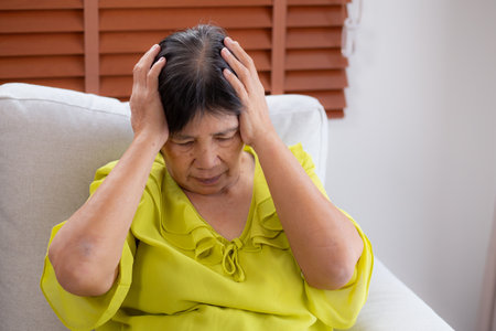 Senior asian woman pain and headache sitting on sofa in living room at home, elderly sitting on couch depression and emotion with stress and migraine, frustration and worried, unhappy and exhaustion.の写真素材