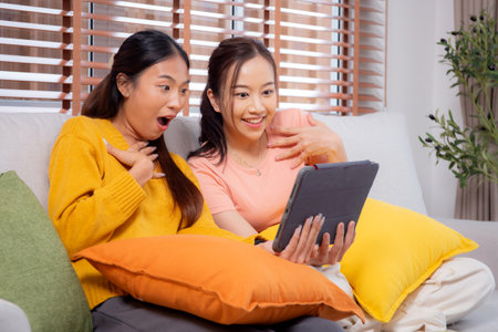 Two young asian woman sitting on sofa using digital tablet for video call in living room at home, female using social media on tablet for online and conversation distant, lifestyles concept.の写真素材