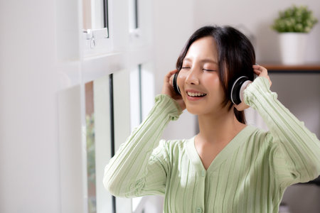 Portrait happy young woman wearing headphones listening music while standing with fun and enjoy in the living room at home, happiness female with entertainment, one person, lifestyle concept.の写真素材