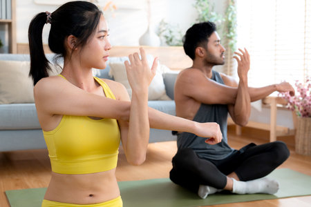 Young asian couple practicing yoga in living room at home, stretching arm pose, fitness and flexibility on yoga mat, man and woman doing yoga together in a cozy home, self-care for health.の写真素材