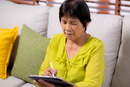 Senior asian woman sitting on sofa using digital tablet with relax in living room at home, elderly sitting on couch writing on tablet with comfortable and leisure, lifestyles and communication.の写真素材