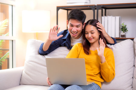 Happy asian couple video call on laptop sitting on sofa in living room at home, smiling and waving during online communication with relax, couple watching laptop, lifestyle and interaction.の写真素材