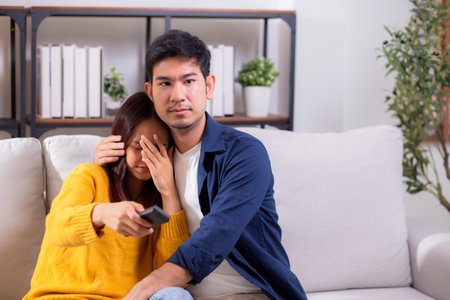 Young asian couple watching a scary or thrilling TV show in living room at home with emotion shock and scared, emotion reaction while watching horror television with fear, lifestyle and bonding.の写真素材