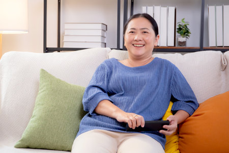 Asian woman sitting on a sofa watching TV with a remote control in living room at home, happy woman relax in a cozy and enjoy entertainment on movie, contentment and lifestyle for fun and satisfied.の写真素材