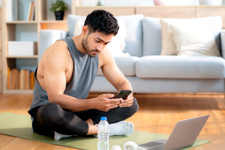 Young asian man sitting on yoga mat relax after home exercise using smartphone, modern healthy and digital lifestyle, fitness routine, man looking smart phone after workout, self-care and wellness.の写真素材