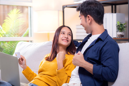 Happy asian couple using laptop and credit card for online shopping together in living room at home, man and woman smiling and relax with enjoy a digital lifestyle, convenient payment.の写真素材