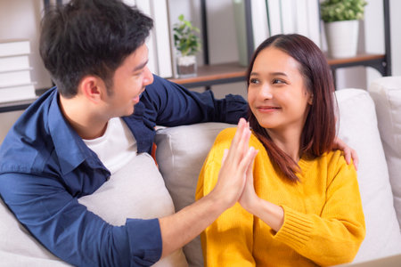 Happy asian couple sitting on sofa in living room at home, celebrating success with a high five while working on a laptop, smiling and enjoying teamwork, remote collaboration, joyful together.の写真素材