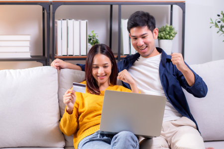 Happy asian couple using laptop and credit card for online shopping together in living room at home, man and woman smiling and relax with enjoy a digital lifestyle, convenient payment.の写真素材
