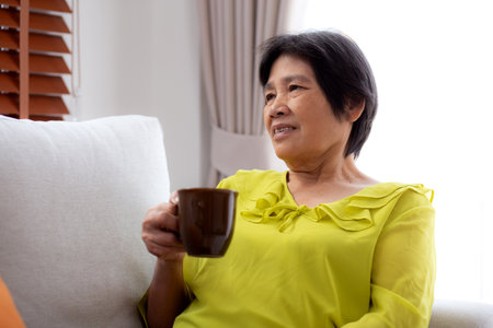 Happiness senior woman sitting on sofa and holding a coffee cup drinking looking out a window in living room at home, elderly drinking beverage for relax, lifestyle and cozy, domestic life.の写真素材