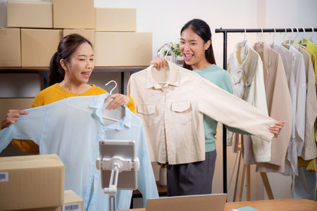 Two young asian woman live online clothing store while showing clothing with tablet on social at home, entrepreneur streaming for selling online clothing and presenting, online seller.の写真素材