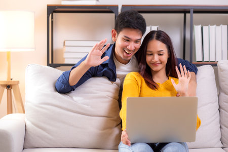 Happy asian couple video call on laptop sitting on sofa in living room at home, smiling and waving during online communication with relax, couple watching laptop, lifestyle and interaction.の写真素材