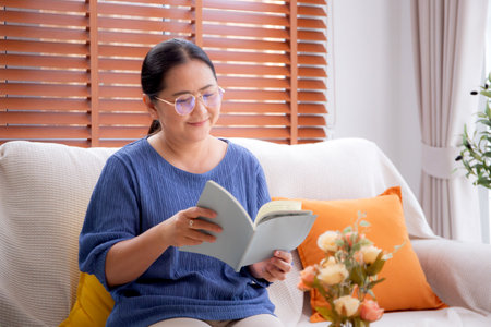 Asian senior woman reading a book sitting on sofa with relax in living room at home, elderly woman reading literature on couch with cozy, hobby and recreation in weekend, lifestyles concept.の写真素材