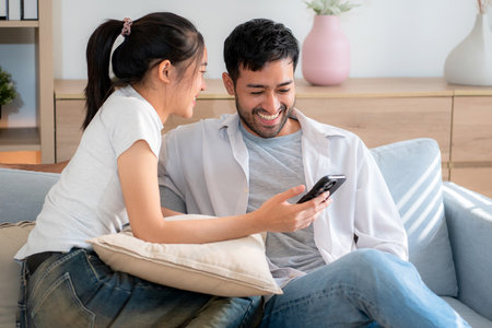 Young asian couple relax sitting on sofa looking smartphone together in living room at home, cozy lifestyle and bonding, relationship and leisure, sharing and enjoy digital content with happy.の写真素材