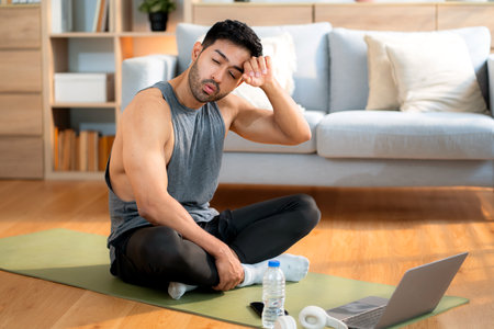Tired young asian man sitting on a yoga mat after a home workout wiping sweat in living room at home, exhausted young man after exercise watching laptop in cozy, self-care and wellbeing.の写真素材