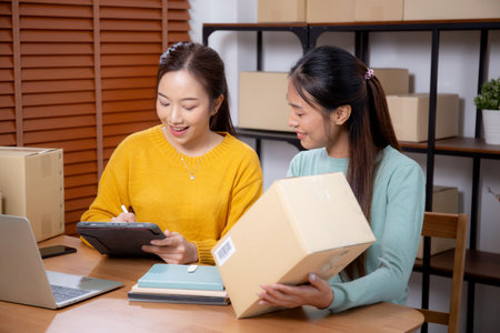 Two young asian woman working while planning online business and checking order with writing on tablet at home, retail with online seller, startup business and cargo, shipping and service.の写真素材