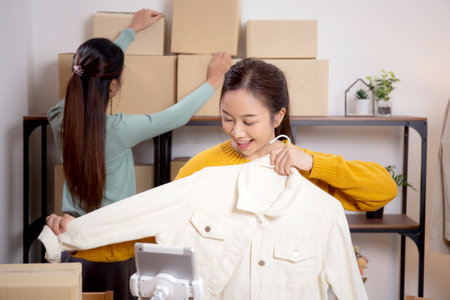 Two young asian woman live online clothing store while showing clothing with tablet on social at home, entrepreneur streaming for selling online clothing and presenting, online seller.の写真素材