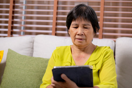 Senior asian woman sitting on sofa using digital tablet with relax in living room at home, elderly sitting on couch writing on tablet with comfortable and leisure, lifestyles and communication.の写真素材
