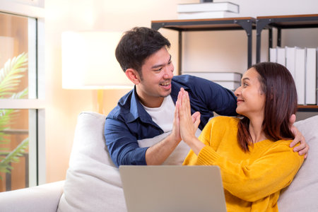 Happy asian couple sitting on sofa in living room at home, celebrating success with a high five while working on a laptop, smiling and enjoying teamwork, remote collaboration, joyful together.の写真素材
