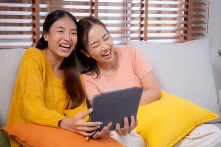 Two young asian woman sitting on sofa using digital tablet for video call in living room at home, female using social media on tablet for online and conversation distant, lifestyles concept.の写真素材