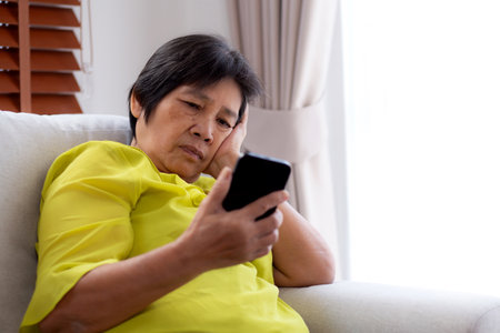 Senior asian woman stress and worried sitting on sofa while looking on smart phone in living room at home, woman unhappy and concerned watching on smartphone, lifestyle and communication.の写真素材