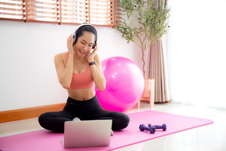 Young asian woman watch laptop after practice yoga and workout and listening music on headphone in living room at home, woman using notebook and listen music after exercise, sport and wellbeing.の写真素材