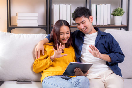 Happy asian couple video call on tablet sitting on sofa in living room at home, smiling and waving during online communication with relax, couple watching tablet, lifestyle and interaction.の写真素材
