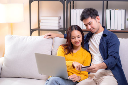 Happy asian couple using laptop and credit card for online shopping together in living room at home, man and woman smiling and relax with enjoy a digital lifestyle, convenient payment.の写真素材