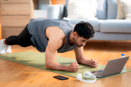 Young asian man doing plank exercise in living room at home while watching online workout tutorial on laptop with endurance and motivation, man training plank on yoga mat, digital fitness lifestyle.の写真素材