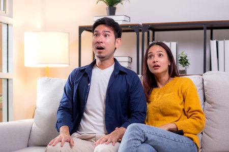 Young asian couple sitting on sofa in living room at home, reacting with surprise while watching...