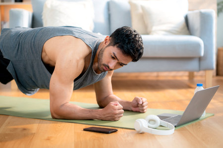 Young asian man doing plank exercise in living room at home while watching online workout tutorial on laptop with endurance and motivation, man training plank on yoga mat, digital fitness lifestyle.の写真素材