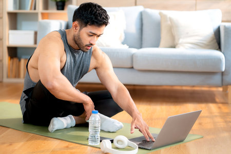 Tired young asian man sitting on a yoga mat after a home workout wiping sweat in living room at home, exhausted young man after exercise watching laptop in cozy, self-care and wellbeing.の写真素材