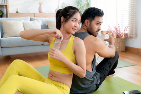 Young asian couple resting together after home workout in living room at home, sitting back to back on yoga mat while drinking water after exercise, refreshment and wellbeing, taking a break.の写真素材