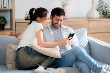 Young asian couple relax sitting on sofa looking smartphone together in living room at home, cozy lifestyle and bonding, relationship and leisure, sharing and enjoy digital content with happy.の写真素材