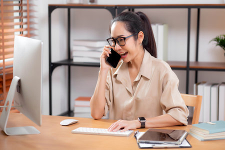 Young asian business woman using desktop computer while talking with smart phone in home office, businesswoman working with desktop computer while speaking with telephone, business or consulting.の写真素材