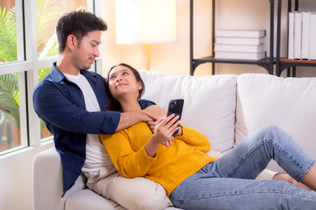 Happy young asian couple relax together in living room at home on cozy sofa while using smartphone, couple embracing using smart phone for resting, affection and lifestyle in modern home.の写真素材