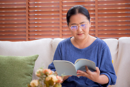 Asian senior woman reading a book sitting on sofa with relax in living room at home, elderly woman reading literature on couch with cozy, hobby and recreation in weekend, lifestyles concept.の写真素材