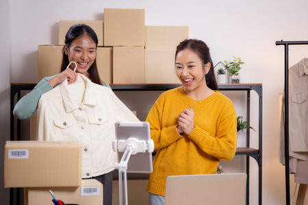Two young asian woman live online clothing store while showing clothing with tablet and laptop on social at home, entrepreneur streaming for selling online clothing and presenting, online seller.の写真素材