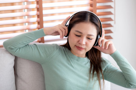 Happy young asian woman wearing headphones listening music while sitting on sofa with fun and enjoy in the living room at home, happiness female with entertainment, one person, lifestyle concept.の写真素材