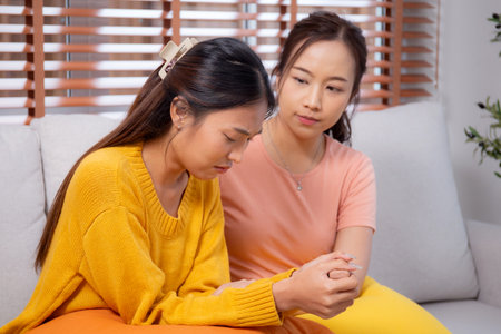 Young asian woman comforting friend with crying and depression sitting on sofa in living room, distress and empathy with friendship, hopelessness and mental health while woman encourage friend.の写真素材