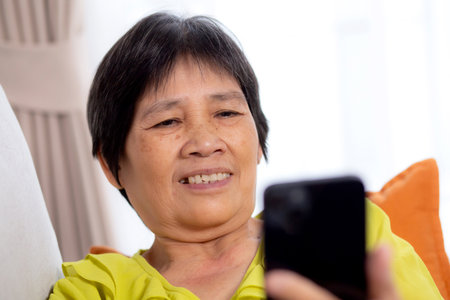 Senior asian woman sitting on sofa looking smartphone for video call in living room at home, elderly using social media on smart phone for chat online and conversation distant, lifestyle concept.の写真素材
