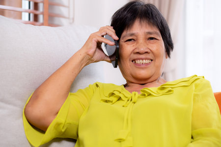 Senior asian woman telephone smiling and relax sitting on sofa in living room at home, cheerful elderly woman talking with smart phone sitting on couch in home, lifestyle and communication.の写真素材