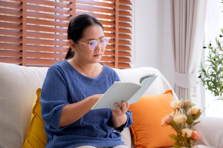 Asian senior woman reading a book sitting on sofa with relax in living room at home, elderly woman reading literature on couch with cozy, hobby and recreation in weekend, lifestyles concept.の写真素材