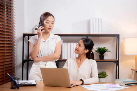 Two young asian businesswoman planning and strategy about marketing and financial while colleagues talking smart phone, meeting and working on laptop computer with partner, business and consulting.の写真素材