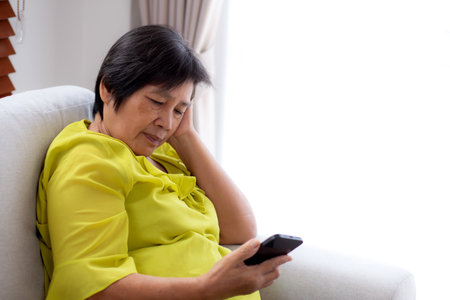 Senior asian woman stress and worried sitting on sofa while looking on smart phone in living room at home, woman unhappy and concerned watching on smartphone, lifestyle and communication.の写真素材