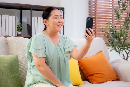 Asian senior woman smiling video call with smartphone in cozy living room at home, elderly sitting on sofa using social media on smart phone, communication and technology, emotion and expression.の写真素材
