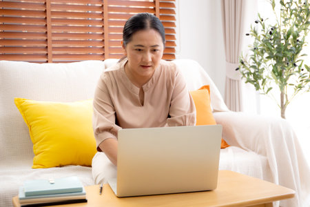 Asian woman working or studying online using a laptop sitting on sofa in living room at home, woman learning for education with determined and inspiration on notebook, e-learning and tutorial.の写真素材