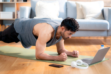 Young asian man doing plank exercise in living room at home while watching online workout tutorial on laptop with endurance and motivation, man training plank on yoga mat, digital fitness lifestyle.の写真素材