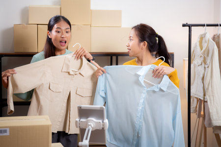 Two young asian woman live online clothing store while showing clothing with tablet on social at home, entrepreneur streaming for selling online clothing and presenting, online seller.の写真素材